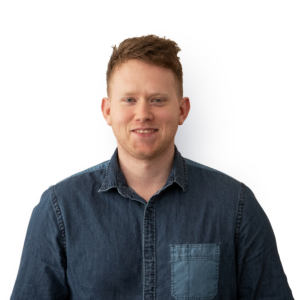 A young man wearing a denim style smart shirt, with short ginger hair and facial stubble