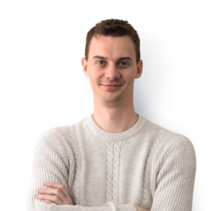 A clean shaven man with short brown hair, wearing a cream jumper with arms folded